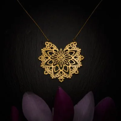 KIWA jewellery brand product image of a large brass pendant necklace called Abundance. The necklace is set on a dark granite background with purple colored flower decoration on the lower end