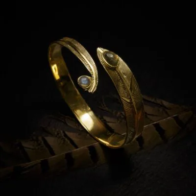 KIWA jewellery brand product image of a single brass armlet called the Feather stone armlet. The armlet has a labradorite stone. The armlet is set on a dark granite background with feather decoration