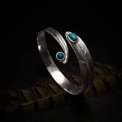 KIWA jewellery brand product image of a single silver plated armlet called the Feather stone armlet. The armlet has a turqoise stone. The armlet is set on a dark granite background with feather decoration