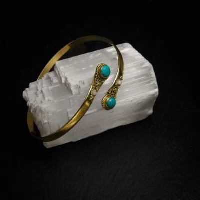 KIWA jewellery brand product image of a single brass armlet called the Four stone armlet. The armlet has a turqoise stone, but it comes in varieties. The armlet is set on a dark granite background on a selenite stone