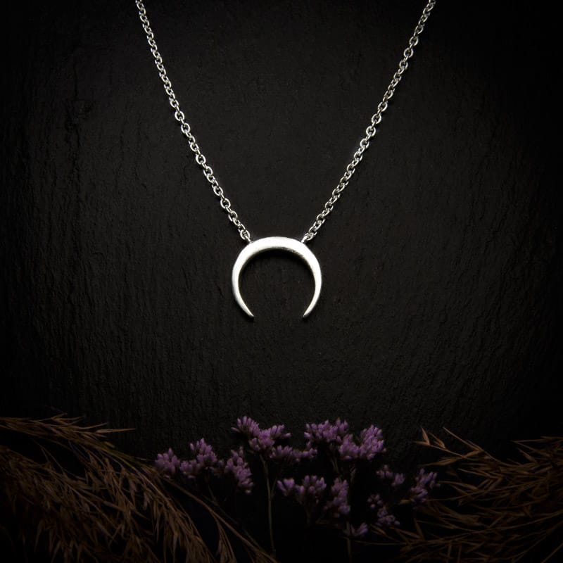 KIWA jewellery brand product image of a silver plated pendant necklace called the New moon. The necklace is set on a dark granite background with hay and dark purple flower decoration on the lower end