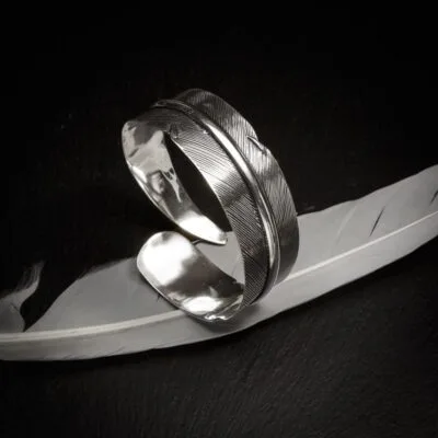 KIWA jewellery brand product image of a single silver plated armlet called the Queen armlet. The armlet is set on a dark granite background with white feather decoration