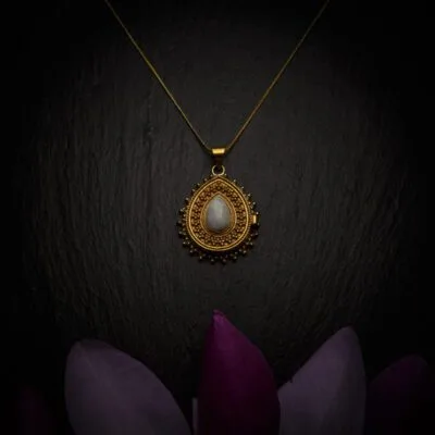 KIWA jewellery brand product image of a brass material pendant necklace called Secret. The necklace has a secret casing inside for hiding valuables and is adorned with a Opalite stone. The necklace is set on a dark granite background with dark purple flower decoration on the lower end