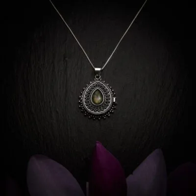 KIWA jewellery brand product image of a silver plated pendant necklace called Secret. The necklace has a secret casing inside for hiding valuables and is adorned with a Labradorite stone. The necklace is set on a dark granite background with dark purple flower decoration on the lower end