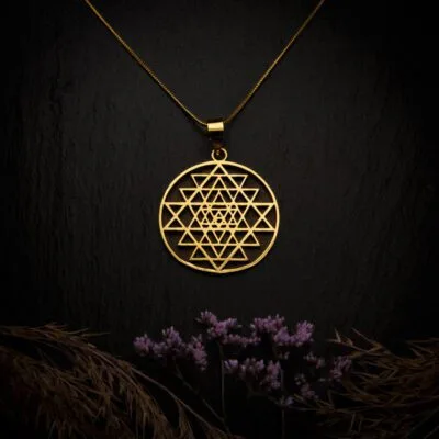 KIWA jewellery brand product image of a single brass material necklace called Sri Yantra, set on a dark granite background with hay and dark purpe flower decoration on the lower end