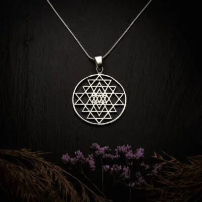 KIWA jewellery brand product image of a silver plated pendant necklace called Sri Yantra. The necklace is set on a dark granite background with hay and dark purple flower decoration on the lower end