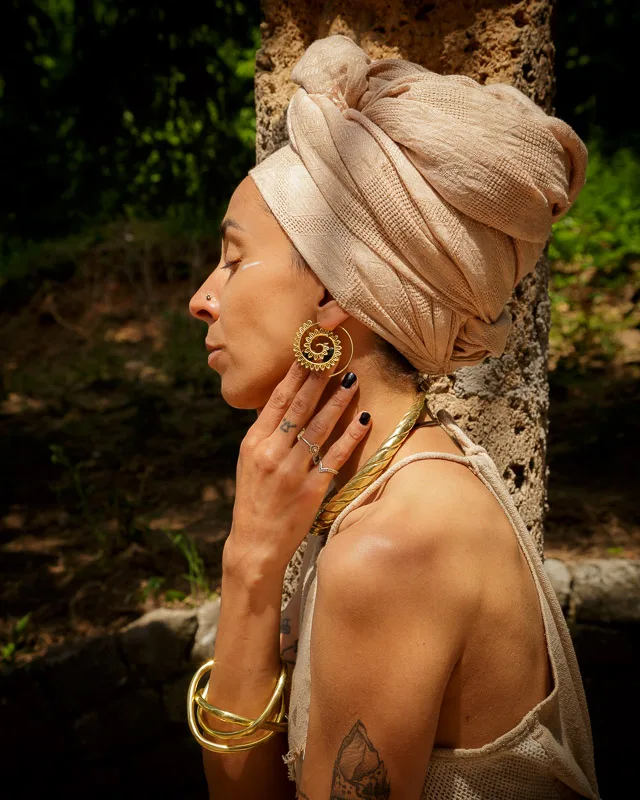 Photo of a Female model wearing Big Spirals Earrings Brass from KIWA Jewellery next to a sand stone pillar in a rosarium in a forest. The model is wearing egyptian style clothing