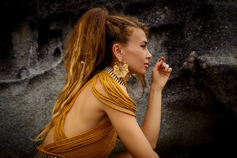 Photo of a Female model Alisa Belochkina wearing Gaia Brass Earrings from KIWA Jewellery handcrafted designs while sitting next to gray beach sand rocks and staring afar