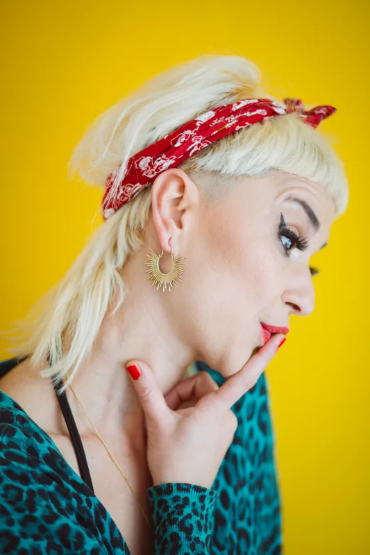 Photo of a Female model wearing Island Earrings Brass from KIWA Jewellery agaist a bright yellow background with a tie around her blonde hair