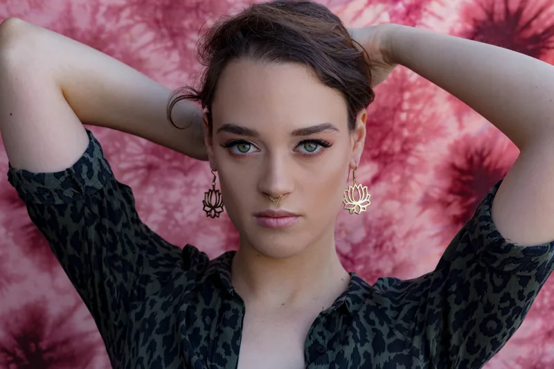 Photo of a Female model wearing Lotus Earrings Brass from KIWA jewelry against a pink background