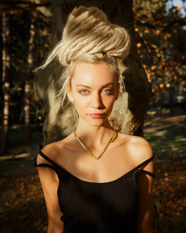 Photo of a Female model Yefimiya wearing Nera Choker Necklace Brass from KIWA Jewellery set against the sun and a platan tree stump. The model has her white dreads all tied up high.