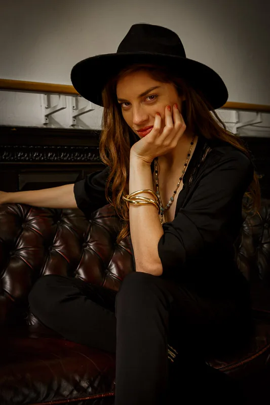 Photo of a Female model wearing Taboo Bracelet Brass from KIWA Jewellery while sitting on an old french style couch in an bohemian apartment while wearing a black hat and staring right at ya