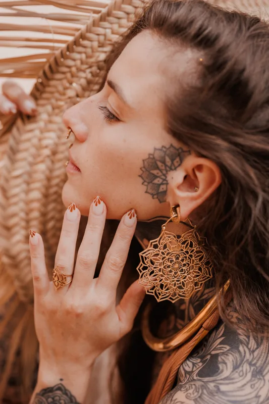 Photo of a Female model wearing Veda Earrings Brass from kiwa jewellery while standing next to a straw hat