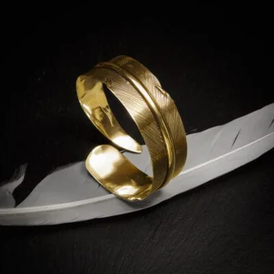 KIWA jewellery brand product image of a single brass armlet called the Queen armlet. The armlet is set on a dark granite background with white feather decoration